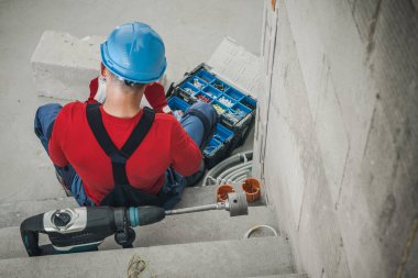 Caucasian Construction Worker Sitting in Front of His Opened Toolbox Preparing to Start Work. Power Drill with Hole Saw Bit Lying Alongside Him. Rear View.