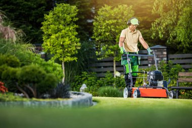 Professional Caucasian Landscaper Wearing Work Uniform and Safety Headphones Mowing the Lawn in the Backyard Landscape Garden Using Petrol Powered Walk Lawnmower. Garden Care and Maintenance Theme.