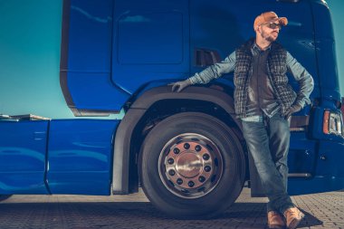 Caucasian Middle Aged Trucker Proudly Standing in Front of His Big Blue Semi-Trailer Truck Looking to the Left Awaiting for the Cargo. Heavy Duty Transportation Theme.