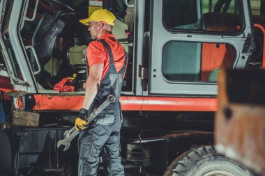 Heavy Duty Machinery Mechanic Standing in Front of Orange Excavator with Large Wrenches in His Hands. Service and Repair Workshop.