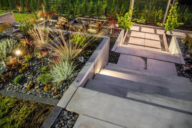 Professionally Landscaped Backyard Garden with Concrete Stairs and Large Tiered Flowerbeds Decorated with Pebbles and Outdoor Lighting Lamps.
