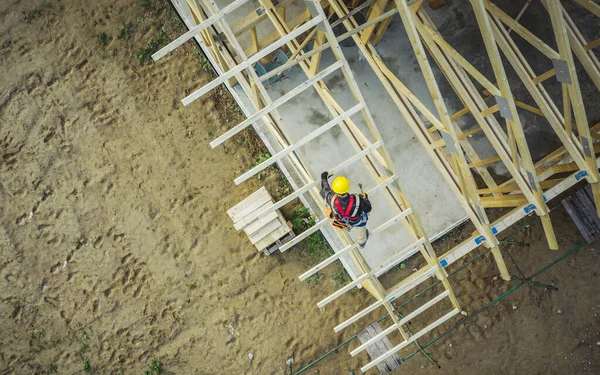 Aerial View of Construction Worker in Yellow Hard Hat Working on Residential Building Wooden Roof Truss to Build a Frame for Roof Covering. Domestic Construction Industry Theme.