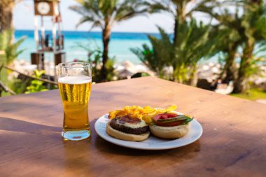 Denize karşı tahta bir masada bir bardak bira ve bir hamburger..