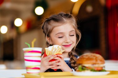 Küçük kız bir fast food Restoran yeme hamburger ve kızarmış patates. Sandviç ve patates cipsi öğle yemeğinde çocuk. Çocuklar sağlıksız yağ yemek yemek. Çocuklar için ızgara fastfood sandviç.