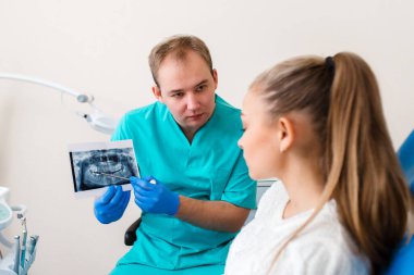 Tıbbi maske takmış, elinde kalem tutan ve dişçilikteki bir hastaya dişlerini gösteren eldivenli esmer bir kadının yan görüntüsü..