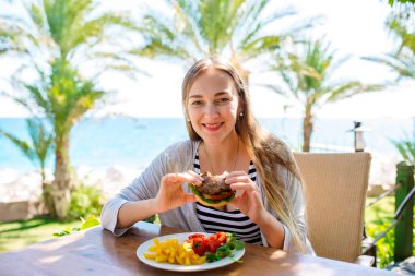 Fast-food, Burgerler, Kahve ve Meyve Suyu İçen Güzel Seyahat Kadını Mavi Deniz ve Arkaplanda Beyaz Kum ile Açık Cafe 'de. Güneşli bir günde Dışarıdaki Restoranda Uzun Saçlı Gülümseyen Kadın