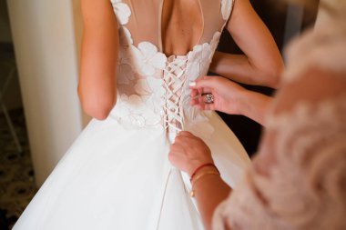 Nedime gelinin beyaz gelinliği iliklemesine yardım eder. Annenin elleri ve düğmeleri gelinin elbisesinde. Gelin gelinliğini giyer ve damatla tanışmaya hazırlanır.