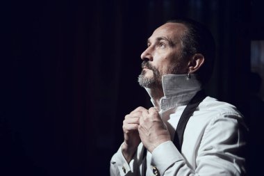 Bir adam tütsülenmiş bir odada kanepede otururken kollarını beyaz bir gömleğe bağlıyor.