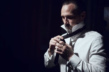 Bir adam siyah bir stüdyoda beyaz bir gömleğe siyah bir kravat takar.