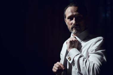 Bir adam siyah bir stüdyoda beyaz bir gömleğe siyah bir kravat takar.