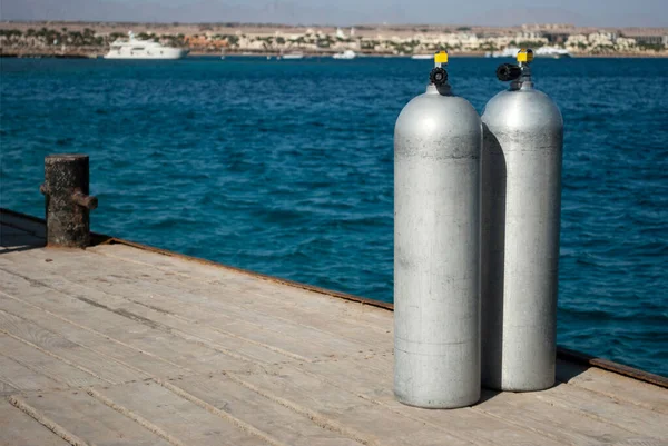 带有氧气的铝气瓶 船坞上有两个潜水员用的钢瓶 码头和大海背景上的全氧气罐蓝色的水和白色的铝圆筒 在码头上关闭两个潜水舱 — 图库照片
