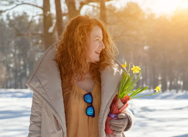 Cheerful red haired woman looking away while walking through winter forest and winter sunshine. charming red haired woman looking away while holding bouquet of yellow narcissus and walking through snowdrifts