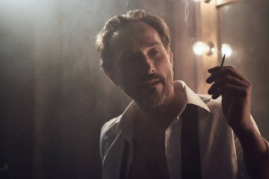 Elinde sigara içen sakallı bir adam. Arka tarafta ışık olan sis odasında bir adam var. Sigara içilen odada beyaz gömlekli bir adamın portresi.