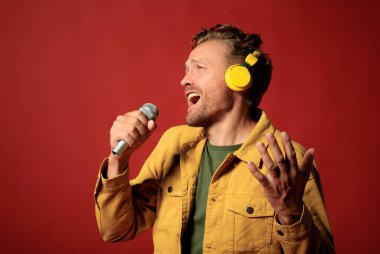 attractive handsome man in yellow jacket and headphones extrimly and emotionaly singing with microphone isolated on dark red background