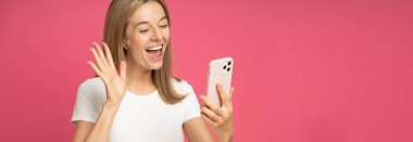 Attractive happy positive woman has video call chatting, waving hand at smartphone camera and smiling, taking selfie, record blog on phone, holding device in hand, say hi, standing on pink background