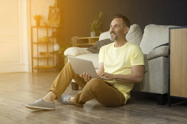 Smiling mature man on floor indoor sitting near of sofa in living room working on laptop compunter Handsome middle-age bearded man looking into sunshine light during his home office hours at home, sitting on a living room floor.