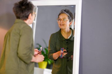 Happy middle aged mature grey haired woman looking in mirrow. Attractive old lady in glasses posing at mirrow.
