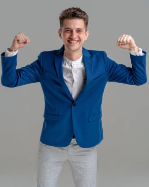 Young smiling happy fun caucasian man 25s in blue jacket and white shirt showing biceps muscles on both hands demonstrating strength power isolated on grey background. happy man showing power in hands. Studio portrait of handsome attractive young man