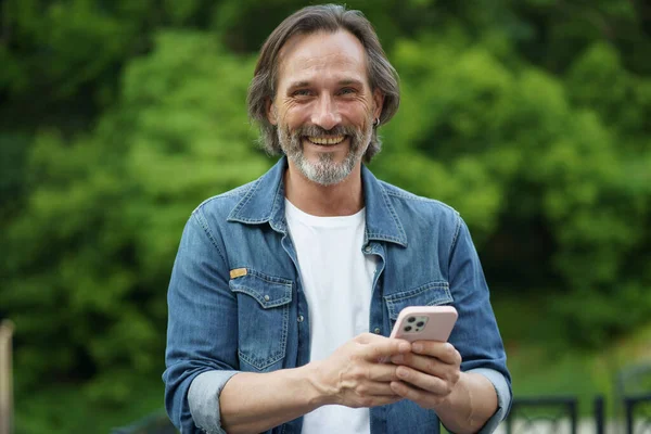 Smiling happy bearded man with mobile phone in hands in green park. Urban mature man in denim shirt walking with phone. Bearded man walking in forest with phone.