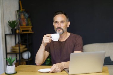 Freelancer evde çalışıyor ve kahve içiyor. Evde bilgisayarla çalışan sıradan tişörtlü sakallı bir adam..
