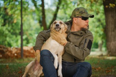 Yeşil ceketli ve şapkalı yakışıklı olgun bir adam ormanda açık havada köpeğe sarılıyor. Parkta köpek gezdiren adam.