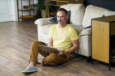 Oturma odasındaki kanepede dizüstü bilgisayarla oturan olgun bir adam..