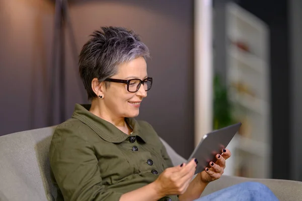 Mature woman reading news at tablet computer sitting on sofa at home. Middle aged woman using pad device while online conversation. Attractive happy woman using internet for communication.