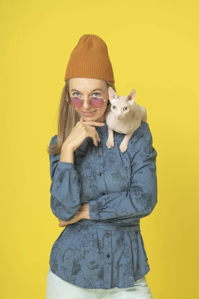 attractive young woman in knitted orange hat with sphynx cat on shoulder. Portrait of cute lovely funny joking attractive woman with white cat looking in camera isolated on yellow background