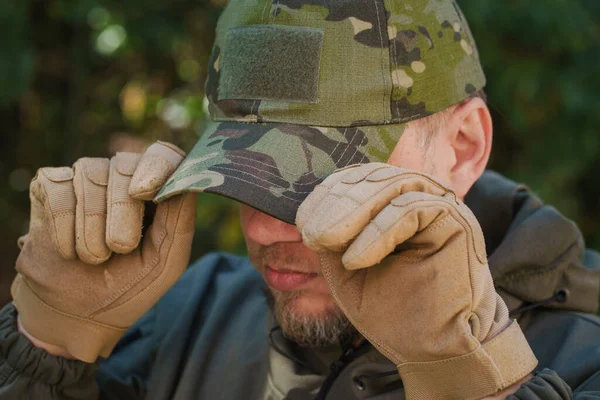 man holds camouflage cap on head by two hands in tactical yellow gloves dressed in dark green camouflage jacket on dark background outdoor. Close up of cap and gloves