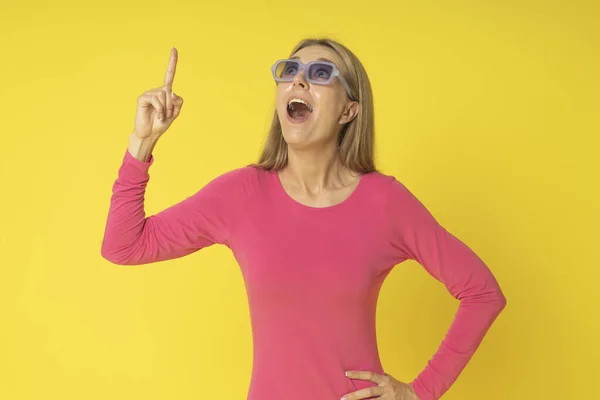Sarı duvar arkasında genç ve çekici bir kadın parmağını kaldırıyor ve gözlüklerle yukarı bakıyor. Heyecanlı, pembe elbiseli, mutlu, sarı elbiseli bir kadın..