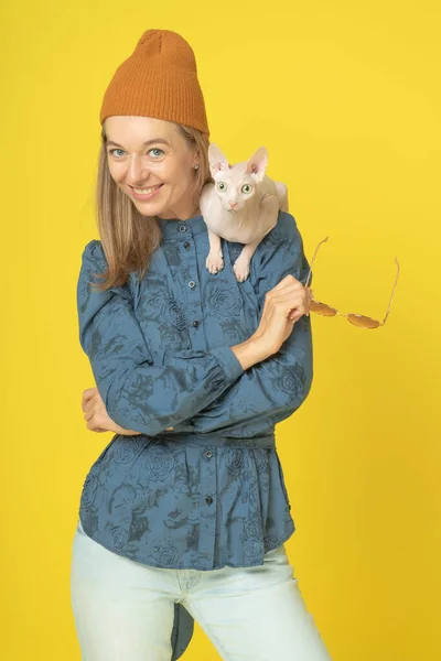 attractive woman in knitted orange hat with canadian white sphynx cat on shoulder. Portrait of cute lovely funny joking attractive woman with white cat isolated on yellow background