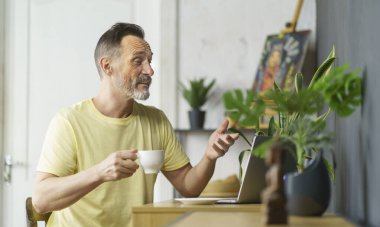 Masada oturan yakışıklı sakallı adam laptopta kahve içiyor videoda sohbet ediyor, erkek çalışan bilgisayar ekranına bakıp el hareketleriyle karar vermeyi düşünüyor.