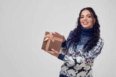 Güzel bir kadın, gri arka plandaki kameraya Noel süveteri giymiş bir hediye kutusu gösterir..