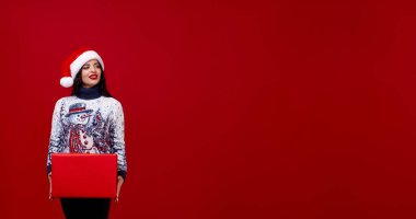 Güzel bir Noel şapkası ve kazağı olan bir kadın kırmızı bir kutu tutuyor ve kırmızı bir arka plana bakıyor. Logosu olan bir hediye kutusu. Yeni yıl afişi. Metin için boşluk.