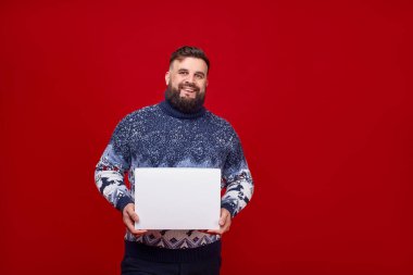 Noel süveteri giymiş gülümseyen bir adam kırmızı arka planda beyaz bir kutu tutuyor. Logo için beyaz bir kutuya tırman. Yeni yılın örgü süveteri. Noel teslimatı.