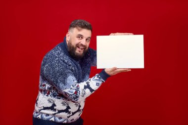 Noel süveteri giymiş sakallı duygusal bir adam kırmızı arka planda beyaz bir kutu gösteriyor. Logo için beyaz bir kutuya tırman. Yeni yılın örgü süveteri.