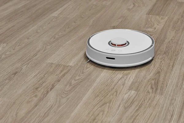 A white robot vacuum cleaner works on a laminate floor. Smart House. The robot vacuum cleaner automatically cleans the apartment.