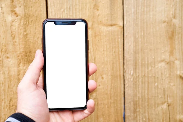 มือของผู้ชายถือโทรศัพท์มือถือกับพื้นหลังของพื้นผิวไม้ รูปแบบโทรศัพท์ที่มีหน้าจอสีขาว — ภาพถ่ายสต็อก