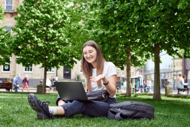 Dizüstü bilgisayarı olan bir kız çimlerin üzerinde oturur ve zoom 'la konuşur. çevrimiçi çalışma