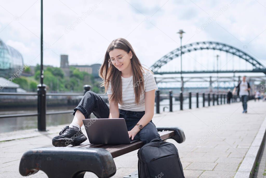 A cheerful girl works with a laptop on the embankment sitting on a bench. 