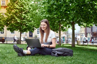 Genç bir kız çimenlerde dizüstü bilgisayarıyla oturuyor.