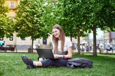Genç bir kız çimenlerde dizüstü bilgisayarla oturuyor. Uzak ofis kavramı..
