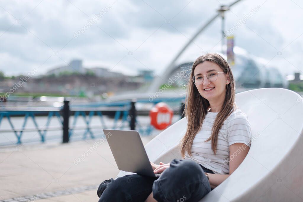 A young woman works at her laptop, sitting on a modern bench on the Newcastle waterfront. Freelancer works on the waterfront. Office without borders.