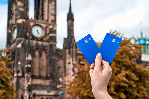 The young womans hand holds blue bank cards against the old chapel building. Bank cards with a chip and contactless payment function. Blue bank cards on a background of autumn trees. Gold autumn.