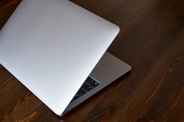 A half-closed laptop with an aluminum lid stands on a wooden table. The keyboard is visible from a half-closed laptop. side view at 45 degrees.
