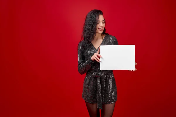 A young girl in a black short dress on a red background holds an empty white box. A beautiful brunette in a black bodysuit holds a white box with space for text or logo. Discount advertising.