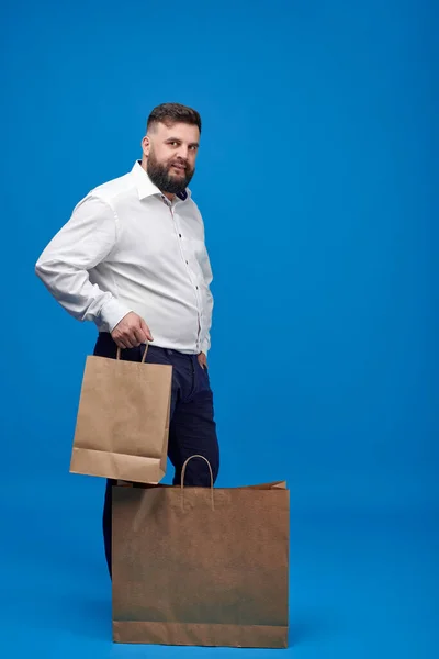 A Caucasian-looking man with a brown paper bag on a blue background. A man with a white shirt with shopping bags. Studio shot of a man with purchases.