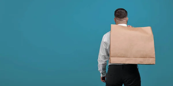 A man with a large brown paper bag on his back stands on a blue background in the studio. Package for close-up purchases. Space for a logo on a paper bag.