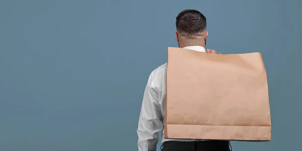 A man with a large brown paper bag on his back stands on a blue background in the studio. Package for close-up purchases. Space for a logo on a paper bag.