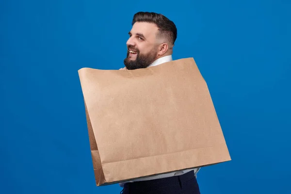 A man with a large brown paper bag on his back stands on a blue background in the studio. Package for close-up purchases. Space for a logo on a paper bag.